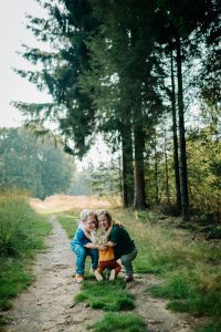 Esther, Tim en Marin geven elkaar een groepsknuffel op een wandelpad in het Sysselt bos in Ede. Je ziet brede glimlachen op de gezichten van Tim en Esther terwijl ze hun blonde dochter samen in de armen sluiten.