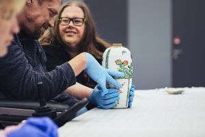 Een deelnemer verkent met handschoenen een kunstobject tijdens een tastworkshop van het Rijksmuseum voor blinden en slechtzienden, terwijl anderen aandachtig meekijken.