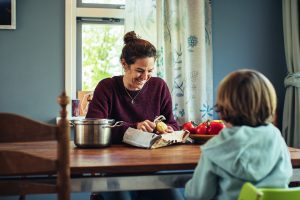 Vrouw schilt biologische groente aan de keukentafel terwijl een kind toekijkt, een rustig moment van duurzaam en bewust leven thuis.