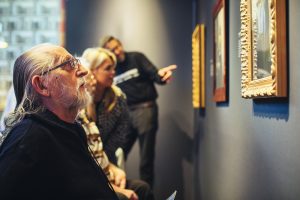 Bewoners van een zorgcentrum bekijken aandachtig schilderijen van Vermeer in museumsetting. Op de voorgrond een een oudere man in rolstoel, zichtbaar onder de indruk en gefocust. Op de achtergrond een medewerker van het Rijksmuseum die de bewoners iets vertelt over de schilderijen.