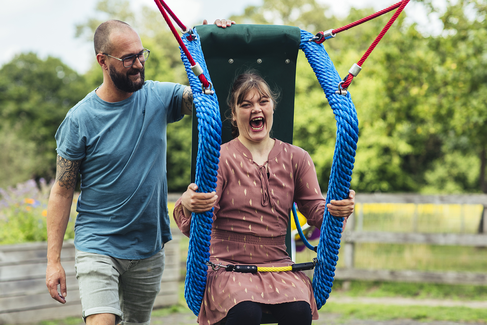 Een jonge vrouw lacht uitbundig terwijl ze op een aangepaste schommel zit in een speeltuin. Een man naast haar duwt haar liefdevol. Vastgelegd moment van plezier dankzij een toegankelijke speelvoorziening, gerealiseerd met steun van HandicapNL.