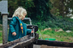 Blond meisje (Marin) speelt met water bij een waterpomp in het Renkums Beekdal, omringd door groen en natuurbeleving.