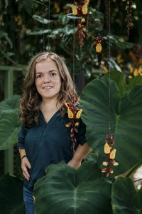 Een portret van Esther Meijer met halflang krullend haar, staand tussen grote tropische bladeren en hangende geel-rode bloemen in de Hortus van Leiden. Ze kijkt glimlachend in de camera en straalt rust en openheid uit te midden van het weelderige groen.
