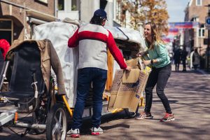 Een vrijwilliger van het Afvalcollectief fietst door de Wageningse binnenstad samen met een nieuwe inwoner. Ze lachen terwijl ze kartonnen dozen opvouwen en in een bakfiets laden. De scène straalt toegankelijkheid, samenwerking en verbinding uit.