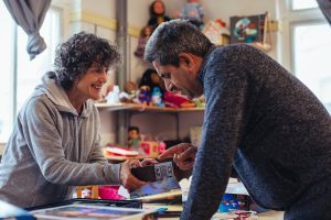Een vrijwilliger van VluchtelingenWerk Nederland helpt een nieuwkomer in een kringloopwinkel. Samen bekijken ze een voorwerp, lachend en in gesprek. De omgeving vol speelgoed en tweedehands spullen onderstreept de praktische, laagdrempelige manier waarop de vrijwilliger ondersteunt bij taal, integratie en wegwijs worden.