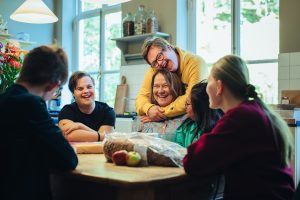 Een groep jongeren zit samen aan een houten keukentafel, ontspannen en lachend. Eén jongen met Downsyndroom omarmt - met een brede lach op zijn gezicht - Linda Germs, die zichtbaar geniet van het moment. Het tafereel straalt warmte, gelijkwaardigheid en plezier uit, in een lichte keukenruimte met grote ramen.