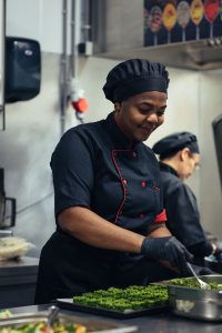 Rebecca, kok uit Liberia, werkt geconcentreerd in de professionele keuken van De Blaauwe Kamer in Wageningen. Ze draagt een zwart kokstenue met rode accenten en schept zorgvuldig fijngehakte groene kruiden in een schaal. Met een rustige glimlach is haar vakmanschap zichtbaar; op de achtergrond werkt een collega, wat de sfeer van samenwerking en ambacht versterkt.