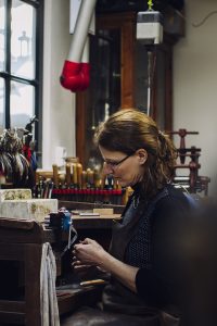 Meestergoudsmid Vivianne Veenemans werkt geconcentreerd aan een sieraad in haar atelier, omringd door gereedschap en materialen. Een plek van ambacht en creatief werk.