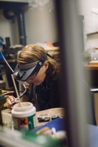 Vivianne Veenemans werkt geconcentreerd aan een sieraad aan haar werkbank, met hoofdlamp en loep, gefotografeerd van een afstand door een raam, alsof je meekijkt in haar creatieve proces.