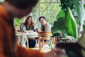 Een vrouw (Linda Germs) zit aan tafel met haar zoon met het syndroom van Down, die een kaartspel vasthoudt, terwijl ze samen opkijken naar een jongen die op de voorgrond piano speelt, in een lichte ruimte met grote ramen en veel groen.