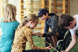 Een groep kinderen staat in een gymzaal dicht om elkaar heen en probeert samen een lange bamboestok langzaam naar beneden te laten zakken met alleen hun wijsvingers, terwijl een jongen in een rolstoel met een glimlach op zijn gezicht meedoet.