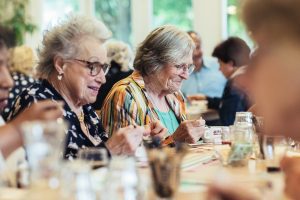 Twee oudere vrouwen zitten samen met buurtgenoten aan lange tafels tijdens een buurtmaaltijd in Wageningen, waar ze samen eten, praten en genieten van elkaars gezelschap in een warme en levendige sfeer.