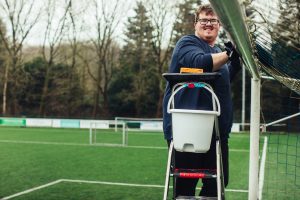 Een jonge man staat op een trap bij een voetbaldoel en maakt met een doek, spons en sop de paal en lat schoon, terwijl naast hem een emmer hangt. Hij kijkt glimlachend naar de camera op het groene voetbalveld.