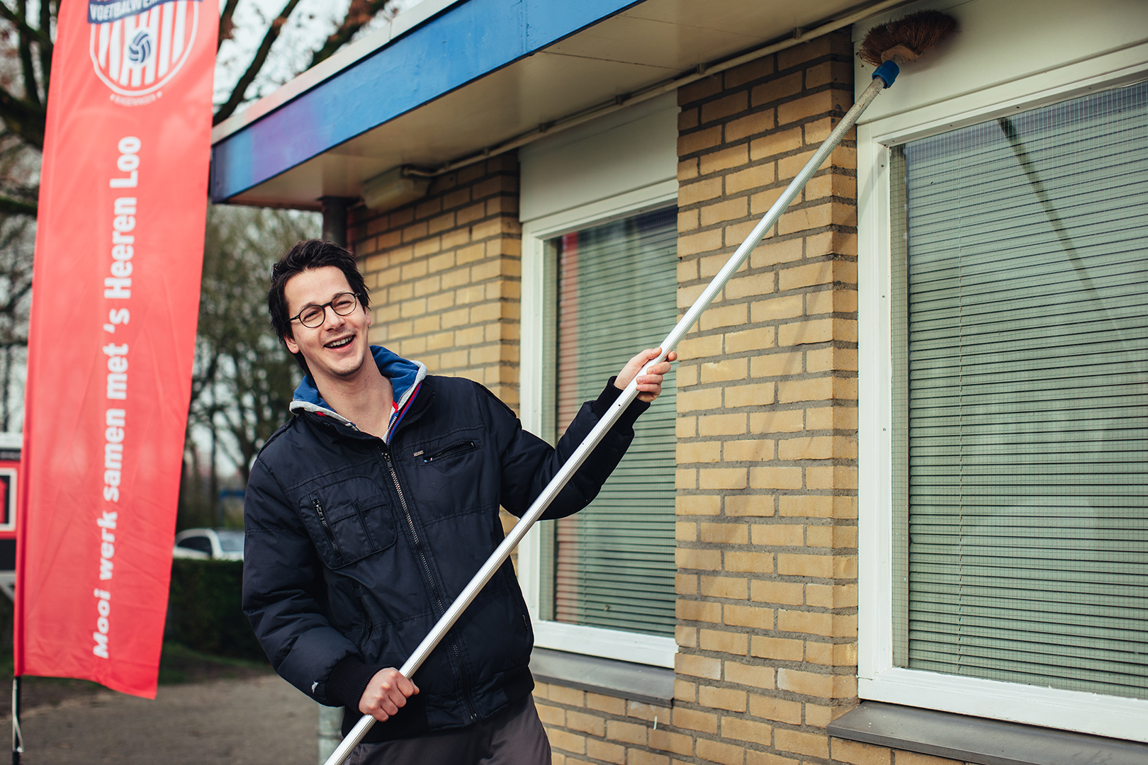 Met een brede glimlach staat een jonge man buiten bij een gebouw en maakt met een bezem de gevel schoon onder de dakgoot, naast een rode banner van de Voetbalwerkplaats met de tekst: “Mooi werk samen met ’s Heeren Loo”.