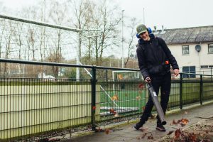Met gehoorbescherming op en een bladblazer in zijn handen blaast een jonge man met een brede lach bladeren over het pad langs het voetbalveld, terwijl de herfstblaadjes opwaaien en zijn plezier in het werk zichtbaar is.