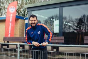Een man in een blauw sportjack met oranje accenten leunt lachend over een hek bij het voetbalveld, met op de achtergrond het clubhuis en een rode banner van de Voetbalwerkplaats met de tekst: “Mooi werk samen met ’s Heeren Loo”.