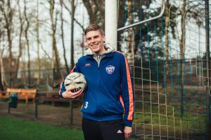 Een jonge man in sportkleding staat ontspannen bij een voetbaldoel en houdt een bal vast, terwijl hij trots glimlachend in de camera kijkt op een voetbalveld van de Voetbalwerkplaats.
