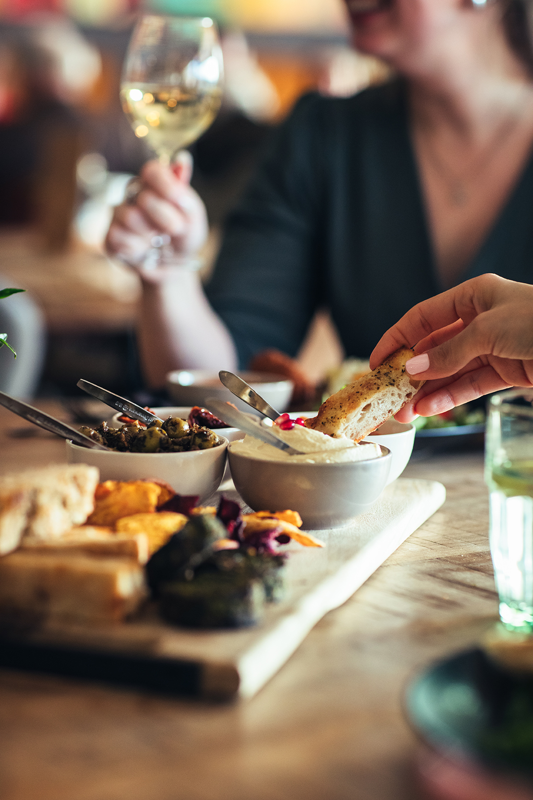 Een hand dipt een stuk brood in een schaaltje hummus op een houten plank met mezze, terwijl op de achtergrond iemand een glas witte wijn vasthoudt. Een sfeervol moment van samen eten, pure ingrediënten en bourgondisch genieten aan tafel.
