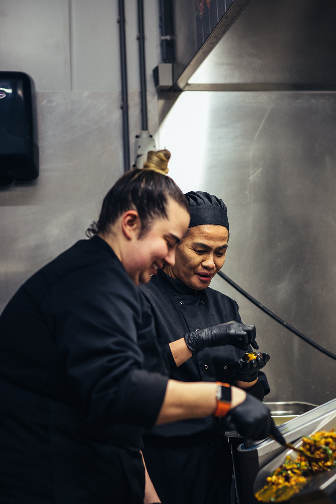 Twee koks in zwarte werkkleding staan samen in een professionele keuken en bereiden met een glimlach op de gezichten een gerecht. Terwijl de ene kok roert en schept proeft haar Thaise collega naast haar het mengsel van groente en bonen. Een moment van samenwerking, vakmanschap en creatief koken in de keuken van een restaurant.