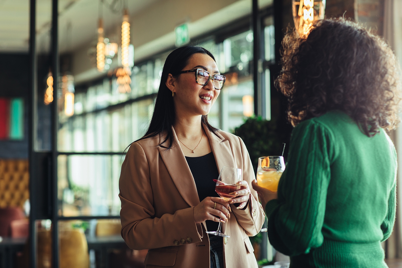 Twee vrouwen staan met een drankje in de hand met elkaar te praten in een sfeervol restaurant met warm licht en grote ramen. Een ontspannen moment van ontmoeting, verbinding en genieten van goed gezelschap in een restaurant.