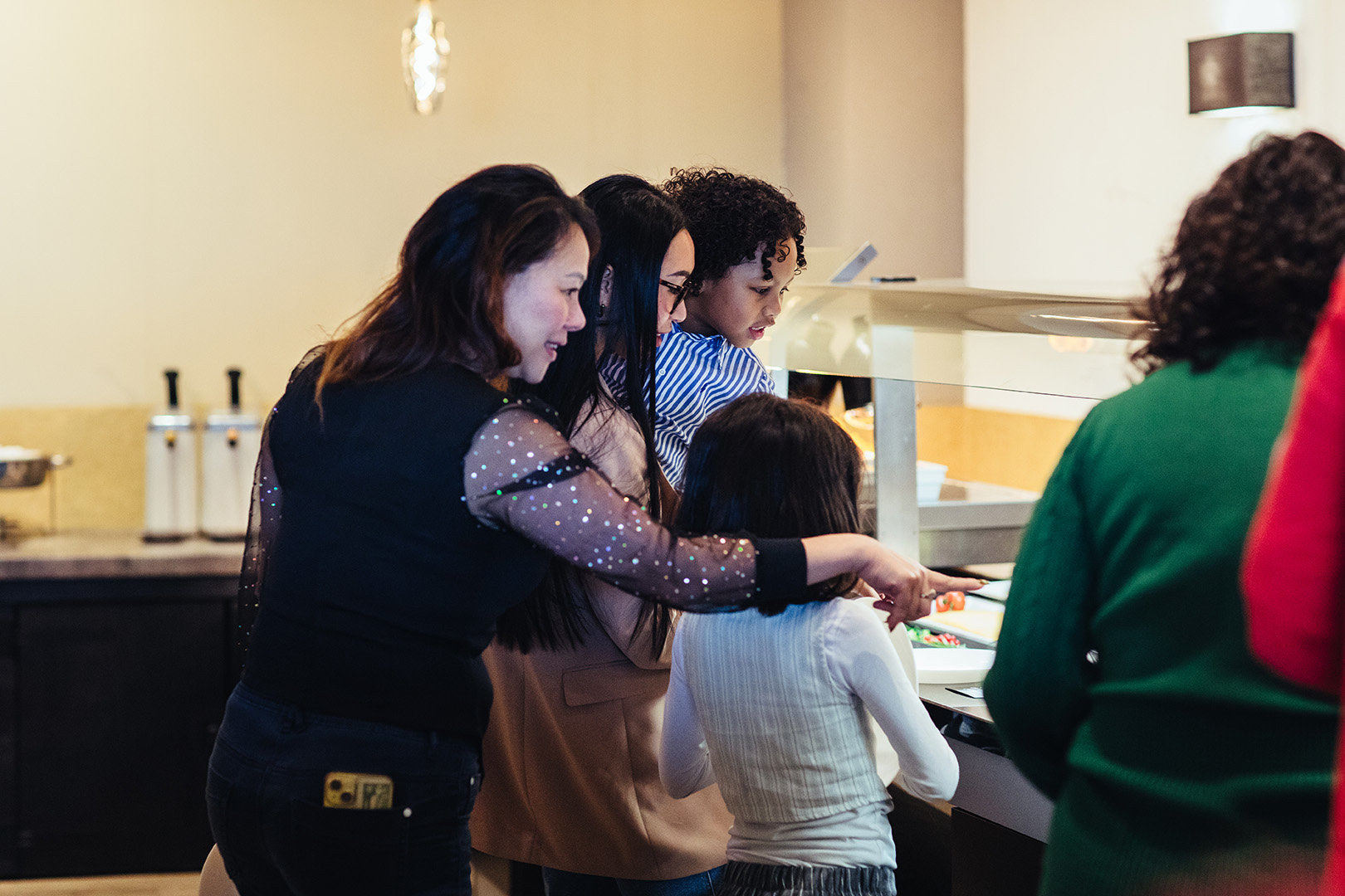 Een groep gasten en kinderen kijkt nieuwsgierig naar het wereldse buffet van De Blaauwe Kamer in Wageningen, terwijl een vrouw glimlachend gerechten aanwijst tussen kleurrijke schalen vol wereldse smaken en warme ontmoetingen.