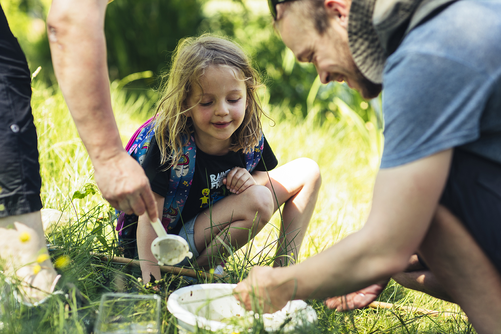 Een meisje zit in het gras en kijkt geboeid toe hoe volwassenen voorzichtig waterdiertjes in een bakje laten glijden. Een rustig, nieuwsgierig moment van ontdekken in het groen.