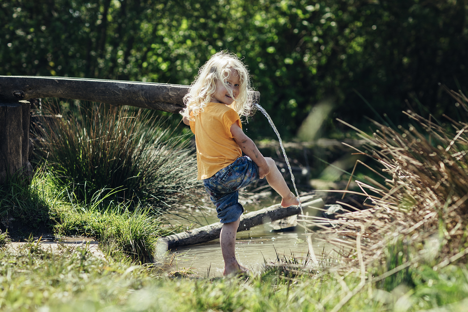 Marin staat lachend met modderige benen en wilde blonde krullen bij de waterspeelplaats in de OERRR Speelnatuur van Natuurmonumenten. Terwijl ze met haar blote voeten in het water speelt tussen het riet, de modder en het zomerse groen van de natuur.