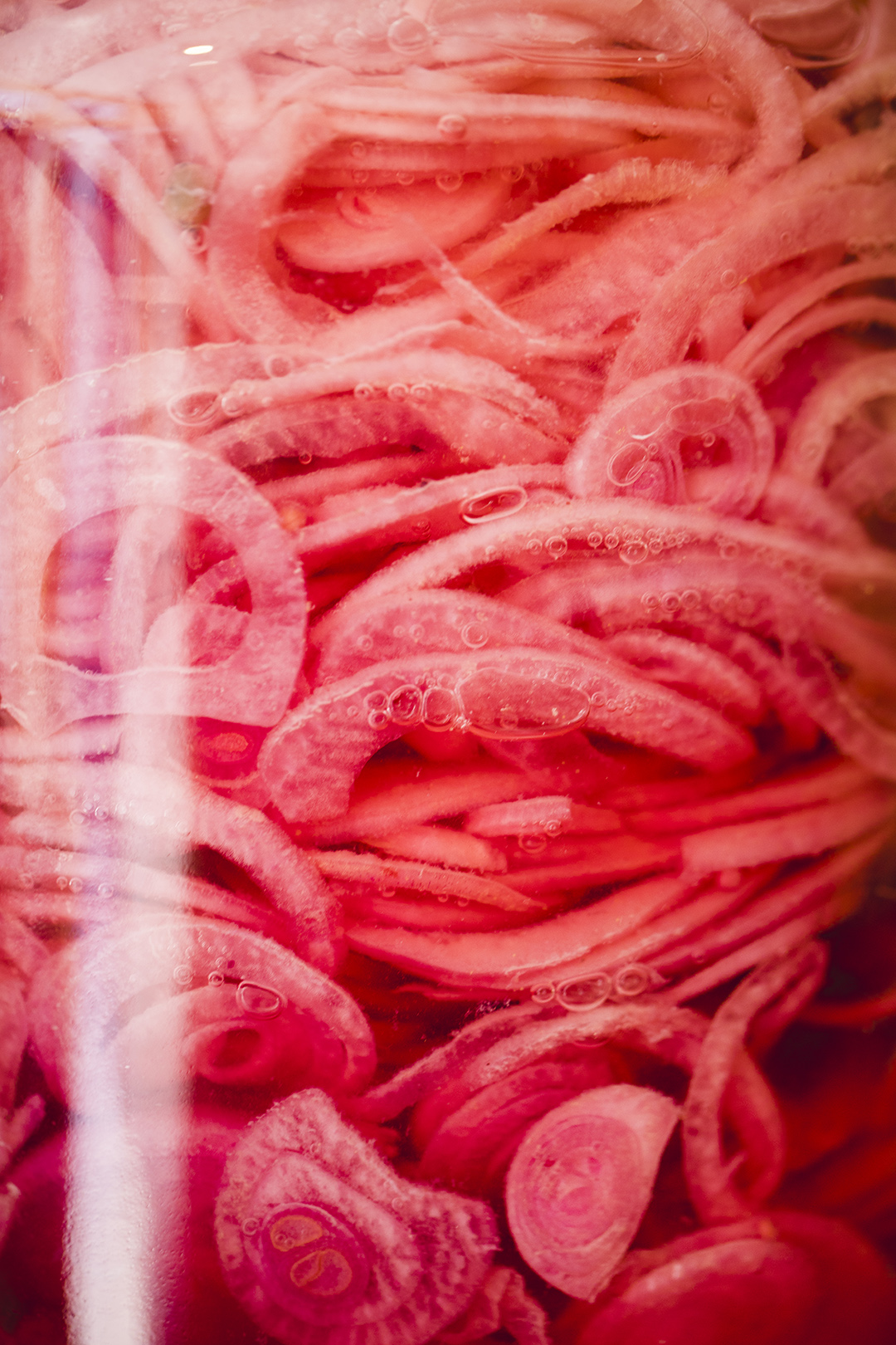 Close-up van dun gesneden rode uien die in een glazen weckpot liggen in zoetzuur, met kleine luchtbelletjes zichtbaar. Het licht weerkaatst zacht op het glas en geeft een dromerig effect. Een sfeervol detail van ambachtelijk inmaken, fermentatie en werken met pure ingrediënten in de keuken.