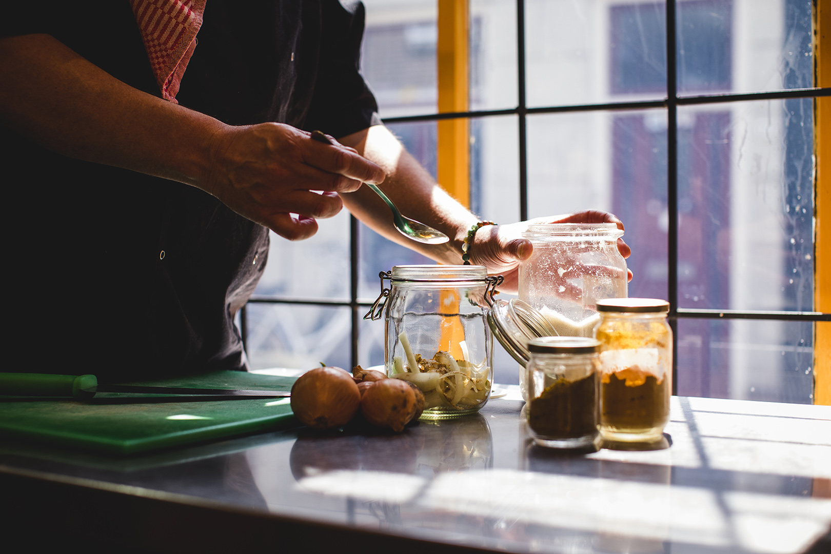 Smaakmaker Chiel Dons voegt met een lepel kruiden toe aan een glazen pot met gesneden uien en specerijen, in een lichte keuken bij het raam. Een sfeervol moment van koken, fermenteren en ambachtelijk werken met pure ingrediënten.