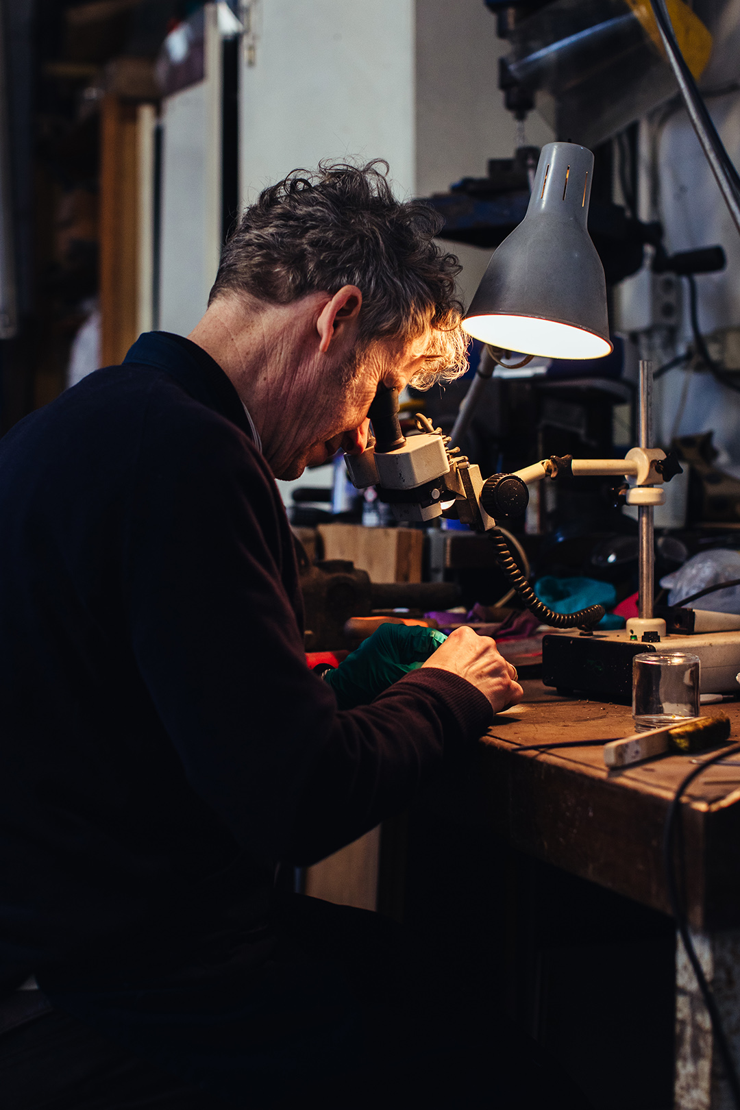Restaurateur Tonny Beentjes werkt geconcentreerd aan een klein object onder een microscoop in een werkplaats, verlicht door een bureaulamp. Een intiem moment van ambacht, precisiewerk en vakmanschap in een rustige werkomgeving.