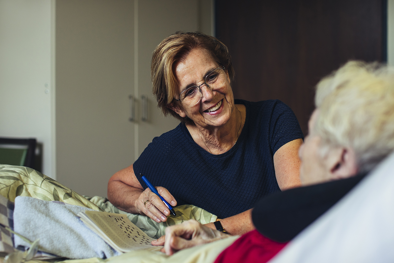Vrijwilliger in een hospice zit naast een oudere vrouw in bed en helpt haar met een puzzelboekje, terwijl ze warm naar elkaar glimlachen. Een intiem moment van palliatieve zorg, aandacht en menselijk contact.