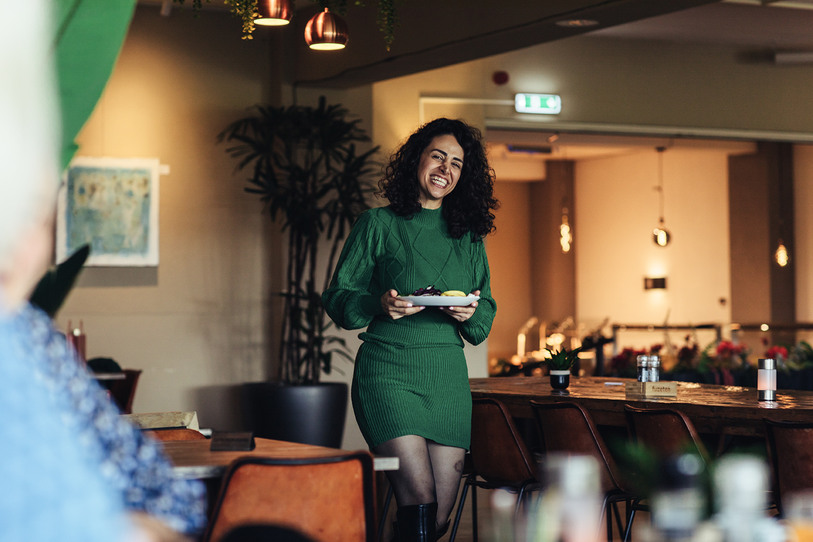Een vrouw met zwarte krullende haren loopt in een groene jurk glimlachend met een bord vol wereldgerechten vanuit de buffetruimte van De Blaauwe Kamer in Wageningen terug naar haar tafelgenoten, die op de voorgrond aan tafel zitten in het warme wereldkeuken restaurant.