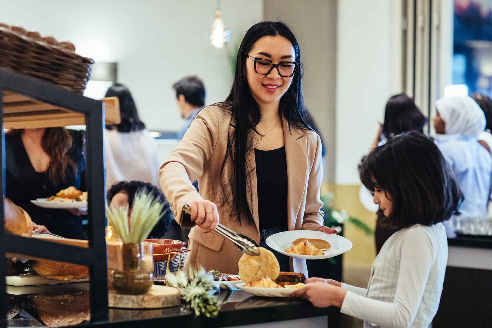 Een vrouw legt glimlachend een kleine American pancake op het bord van een kind bij het wereldse buffet van De Blaauwe Kamer in Wageningen, terwijl ze zelf een bord vasthoudt tussen een levendig en gemêleerd gezelschap van gasten die samen genieten van wereldse gerechten.