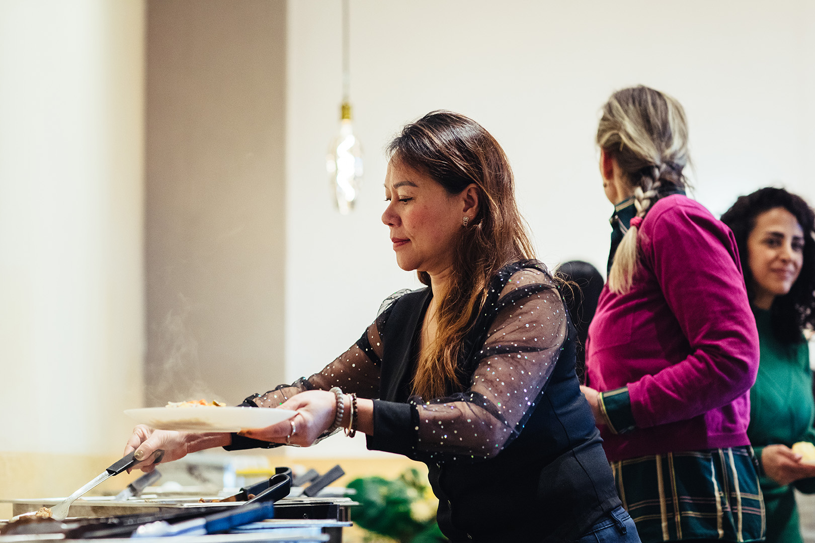Een vrouw schept een warm gerecht op van het wereldse buffet bij De Blaauwe Kamer in Wageningen, terwijl andere gasten op de achtergrond wachten tussen geurige dampen, warme verlichting en een sfeervolle mix van internationale gerechten en ontmoetingen.