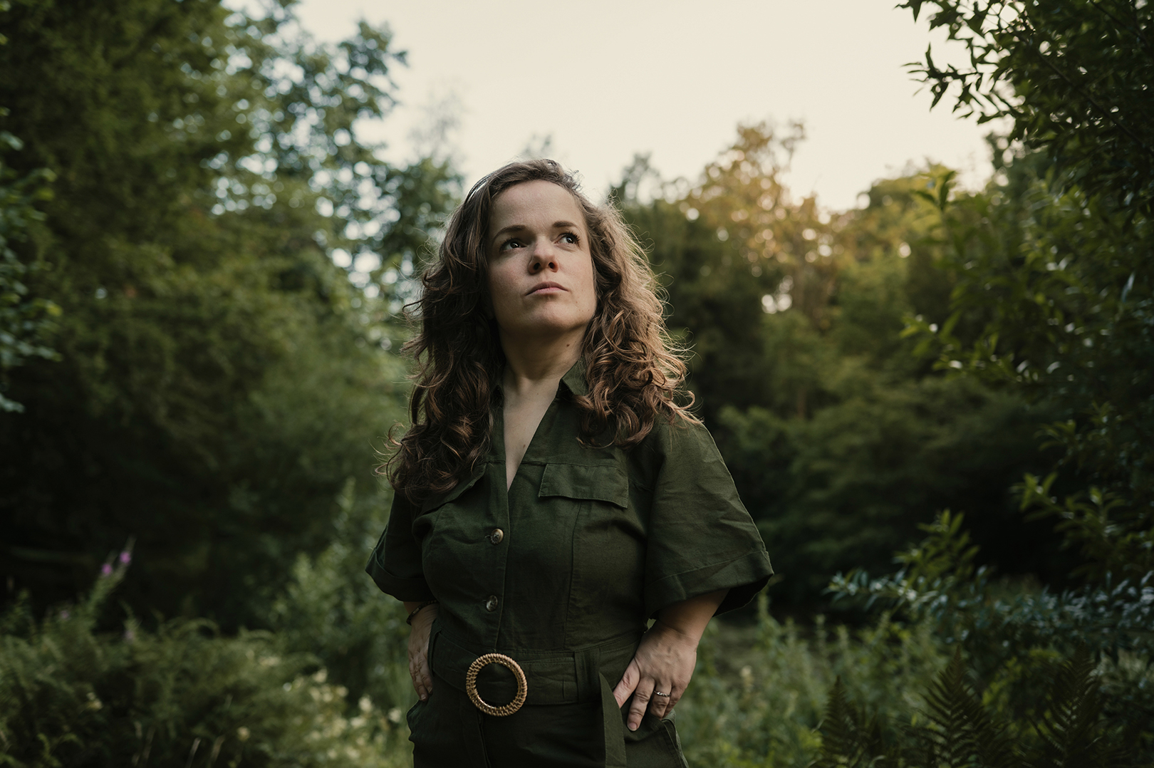 Esther staat tussen de struiken en bomen en kijkt omhoog, alsof iets in de boomtoppen haar aandacht trekt. Haar donkere groene jumpsuit gaat bijna op in het omringende groen, terwijl het zachte avondlicht tussen de bladeren doorschemert.