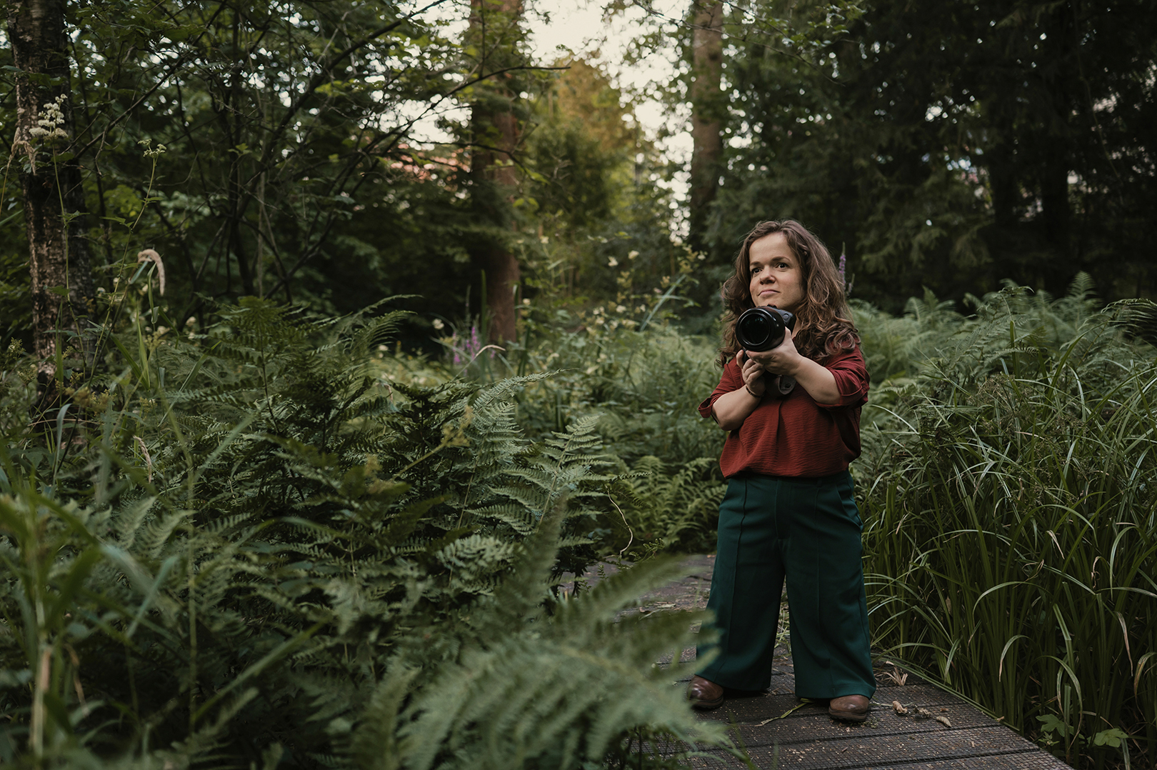 Esther staat met haar camera in haar handen op een houten pad, omringd door hoge varens, struiken en bomen. Haar roestbruine blouse vormt een warm kleuraccent in het uitbundige groen. De ruime compositie laat zien hoe klein ze lijkt te midden van de natuur waarin ze zich zo thuis voelt.