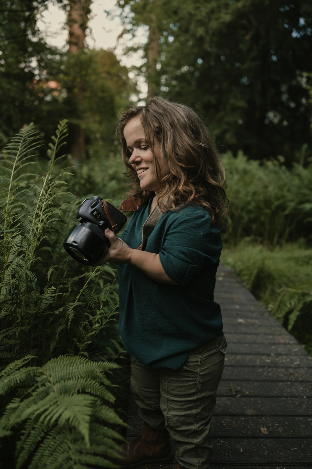 Esther staat op een smal houten vlonderpad, omringd door weelderige varens en frisgroene planten. Met haar camera in de hand kijkt ze geconcentreerd naar wat ze wil vastleggen.