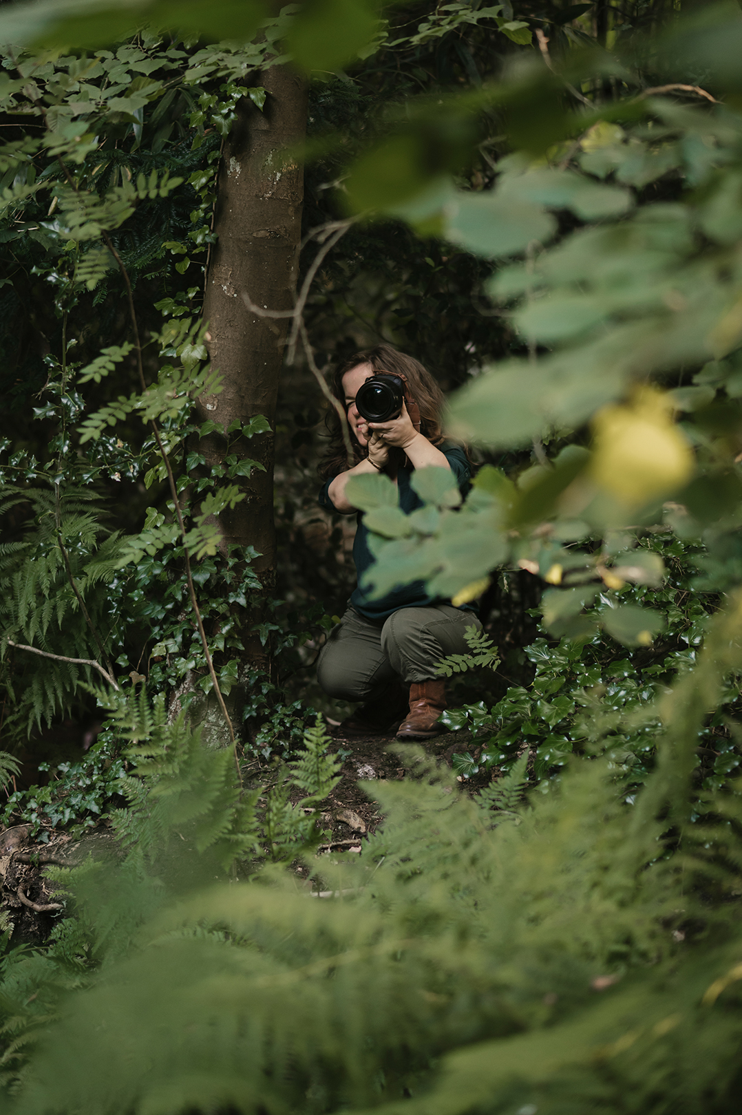 Tussen de bladeren en varens is Esther met haar camera gedeeltelijk verscholen in het groen. Je ziet haar gehurkt tussen de planten, terwijl bladeren op de voorgrond het beeld omlijsten. Alsof je even stiekem meekijkt tijdens het fotograferen in de natuur.
