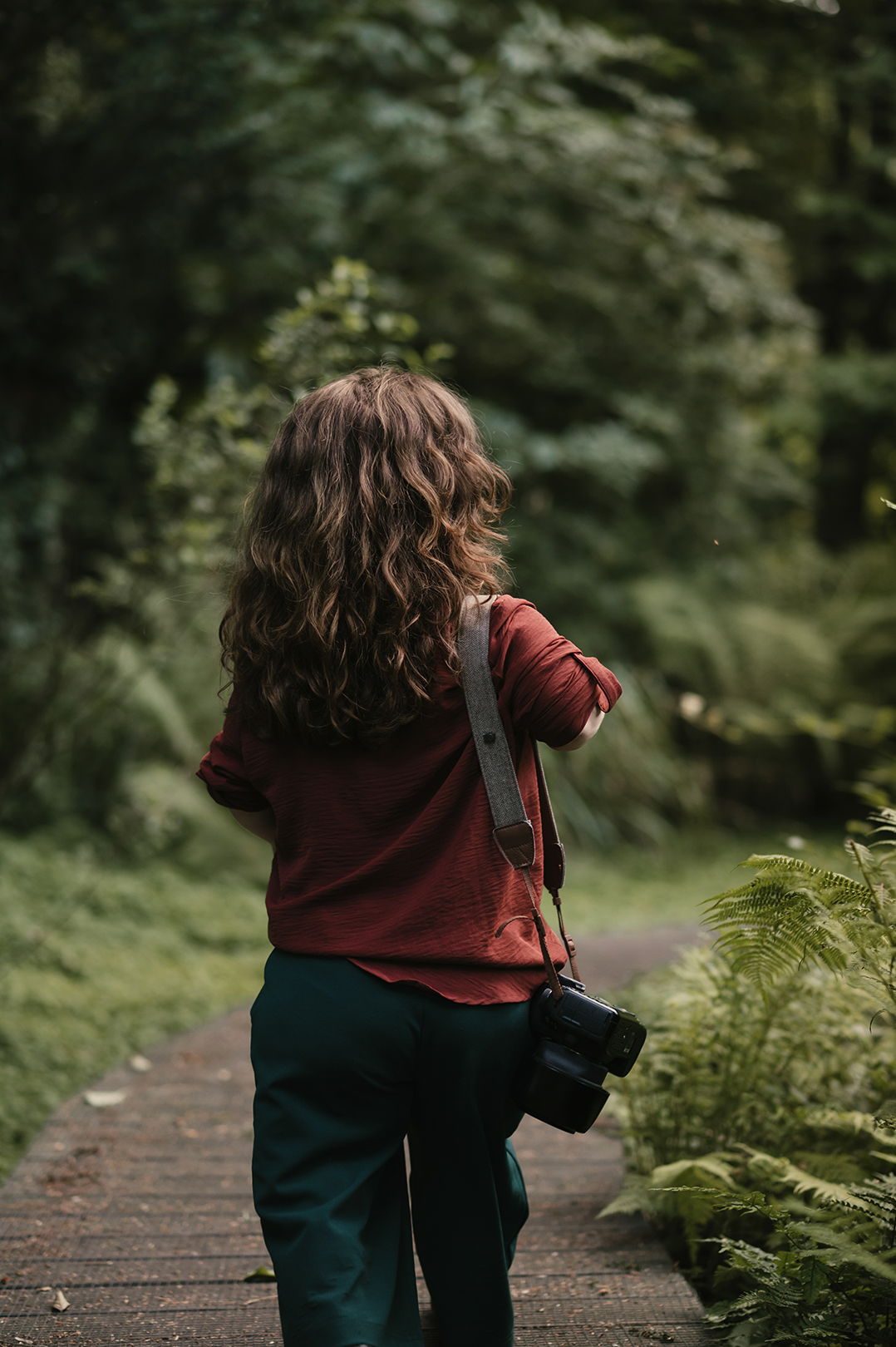 Van achteren zie je Esther over een smal houten pad door het bos wandelen. Haar roestbruine blouse steekt af tegen het diepe groen van de bomen en varens, terwijl haar camera aan haar zijde bungelt.