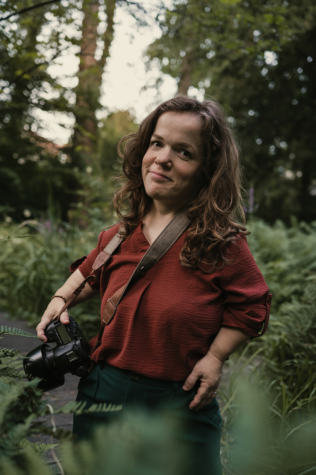 Esther Meijer staat tussen het hoge groen in het bos, met een camera over haar schouder. Ze draagt een roestbruine blouse en een donkergroene broek met wijde pijpen, en kijkt met een ontspannen glimlach naar de fotograaf.