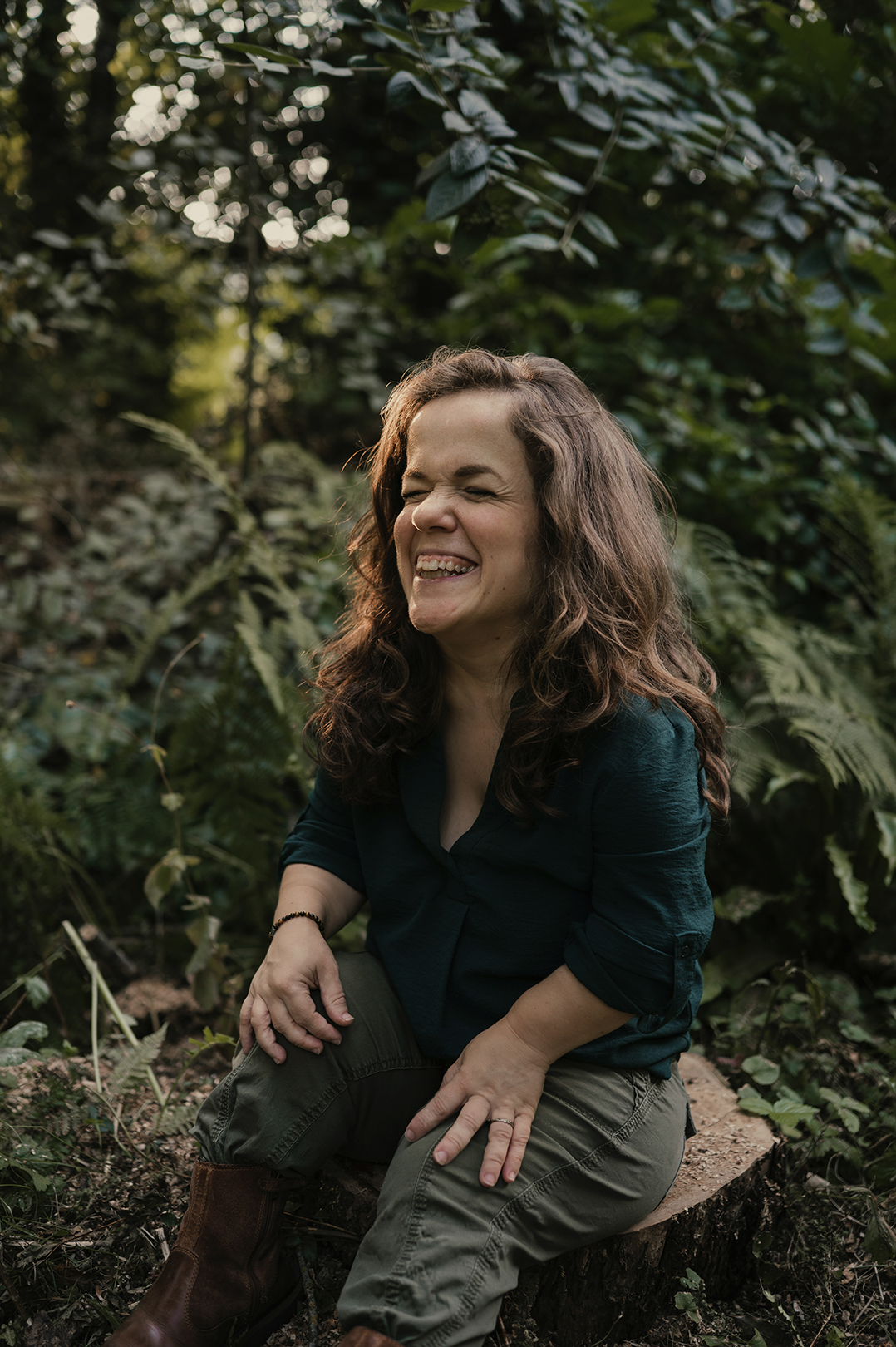 Esther zit ontspannen op een boomstronk tussen het bosgroen en de varens. Ze leunt iets naar voren en lacht breeduit met haar ogen gesloten, en haar krullende haar los om haar gezicht. De foto ademt plezier en ongedwongenheid.