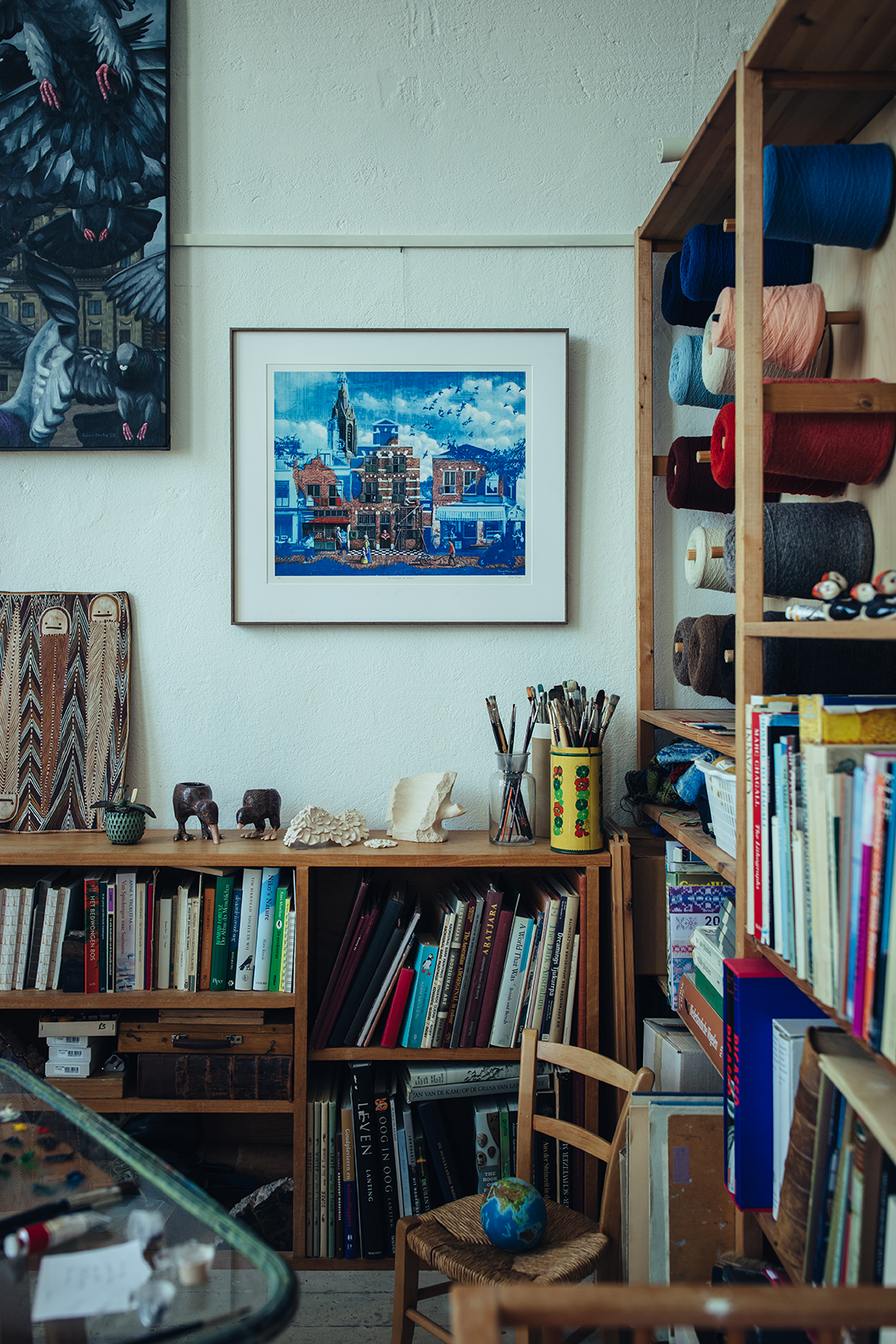 Een kijkje in Rafaels atelier met een ingelijst blauwdruk schilderij van een Delfts straatje aan de muur, boeken en materialen in open kasten, rollen garen en schildersezels. Elke plank en hoek lijkt iets te vertellen over een ander creatief project.