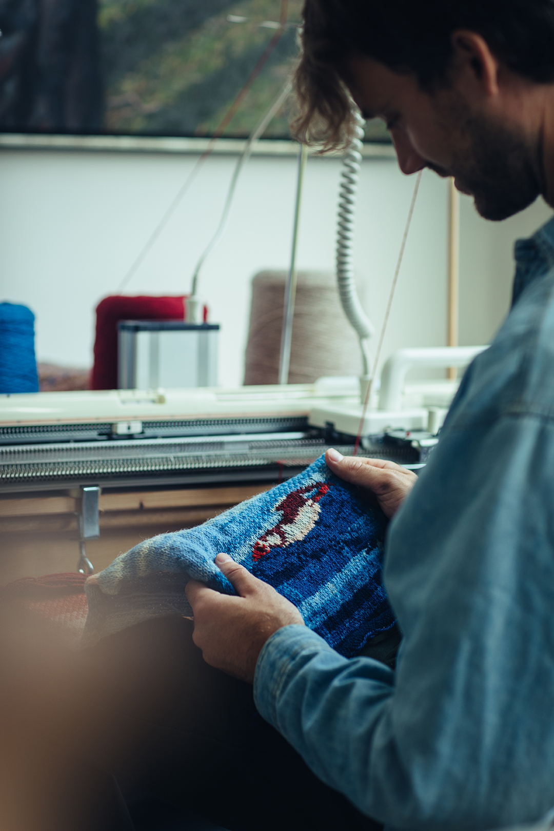Rafael bekijkt een blauw breiwerk waarop een papegaaiduiker is afgebeeld. De kleine steken vormen een fijn reliëf in de stof. Het invallende licht strijkt over de blauwe draden en laat de structuur en het patroon van het breiwerk mooi naar voren komen.