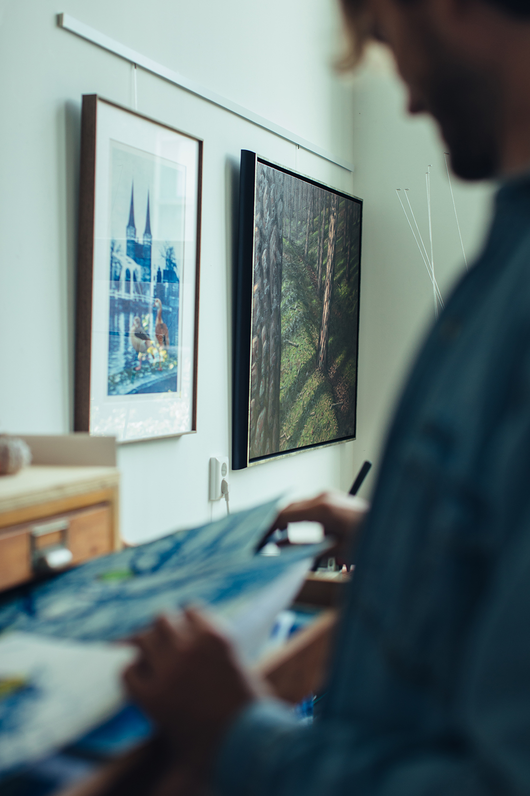 Op de voorgrond bladert Rafael door een lade met blauwe cyanotype werk. Aan de witte muur er achter hangen ingelijste werken van een blauwdruk schilderij met ganzen en een schilderij van een boslandschap, met bomen, mos en diverse schaduwen.