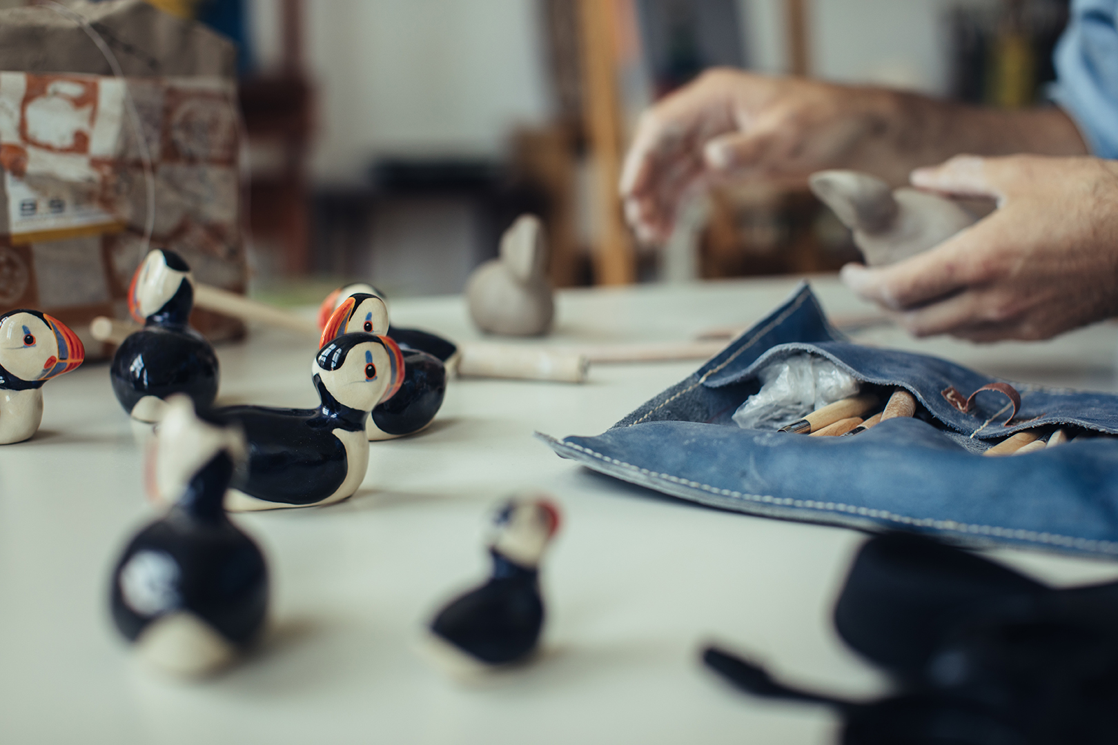 Meerdere kleine keramische papegaaiduikers staan verspreid over een lichte werktafel. Hun zwarte ruggen, witte buikjes en oranje snavels zijn met verf aangebracht op de gebakken klei.