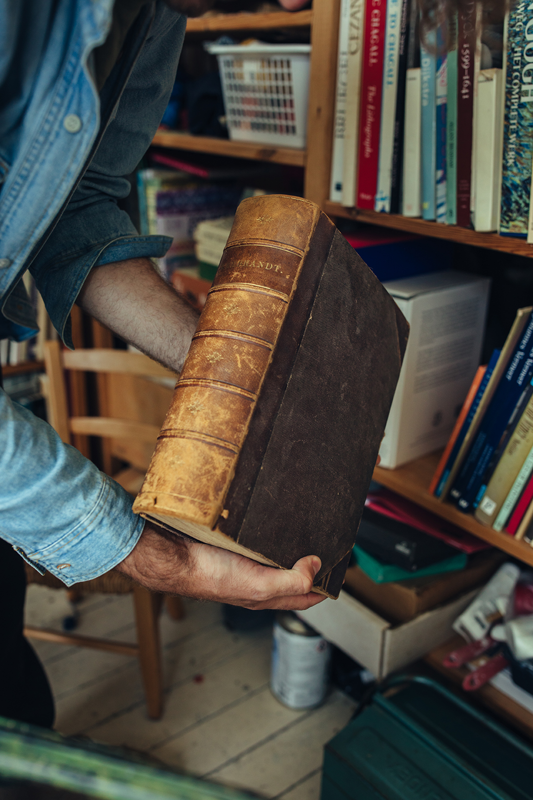 Rafael haalt een oud, bruin boek met een versleten leren kaft uit een volle boekenkast. De verweerde rug met daarop ‘Rembrandt’ in gouden letters, de verkleurde randen en de zichtbare gebruikssporen geven het boek een geschiedenis.