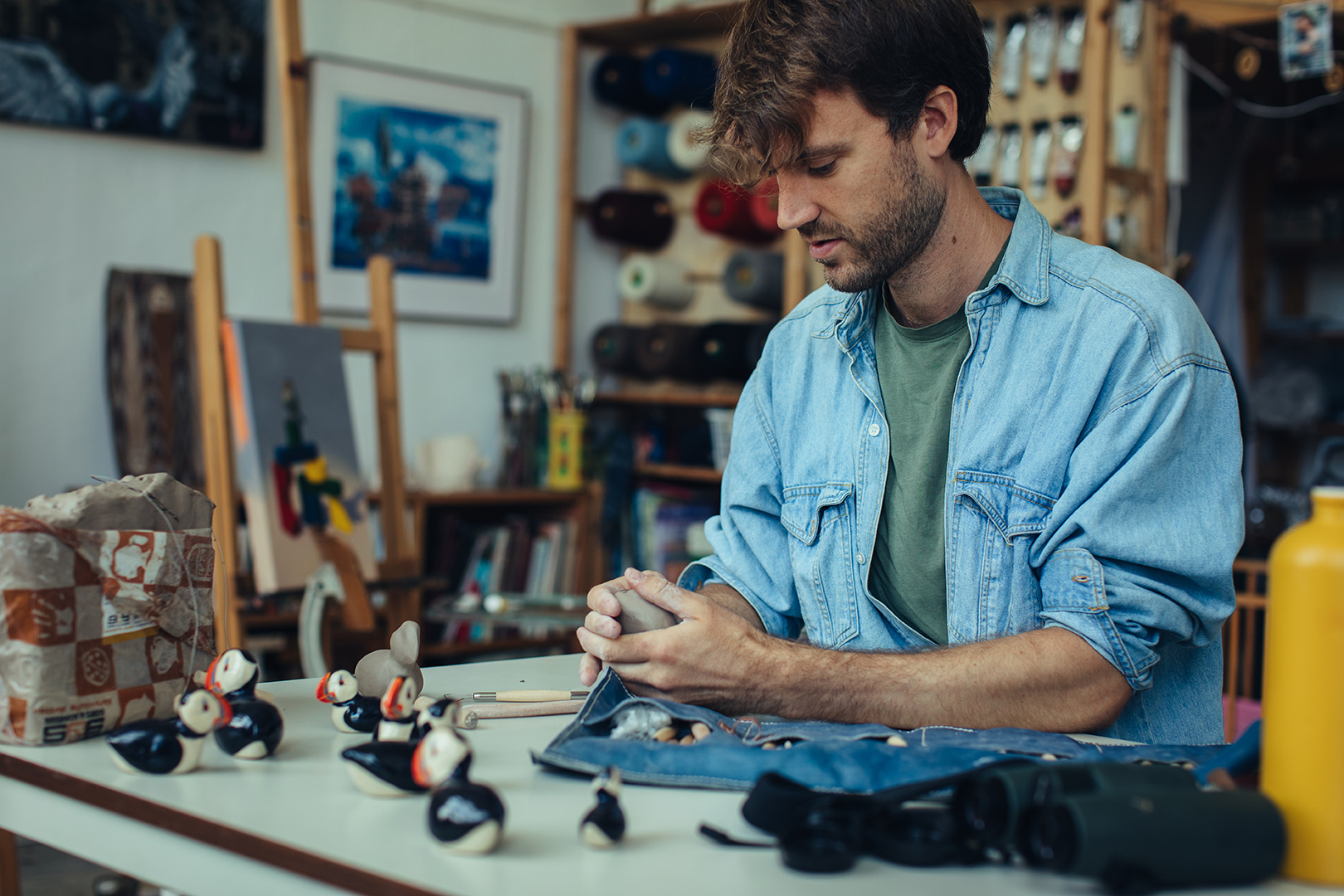 Rafael boetseert aan zijn werktafel met zijn handen een kleine papegaaiduiker uit klei. Hij vormt het lijfje en de contouren van de vogel zorgvuldig met zijn vingers. Om hem heen liggen materialen en andere creatieve projecten in zijn atelier.