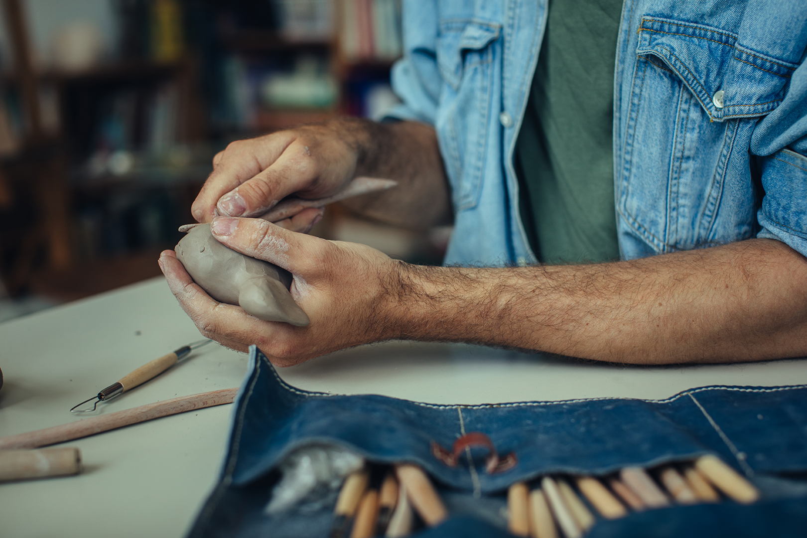 Rafael geeft een kleine papegaaiduiker vorm uit klei met behulp van een houten modelleerstokje. Op de voorgrond ligt een opengerold etui van blauwe spijkerstof met daarin zijn gereedschap. Met het houten instrument brengt hij zorgvuldig details en lijnen aan in de zachte klei.
