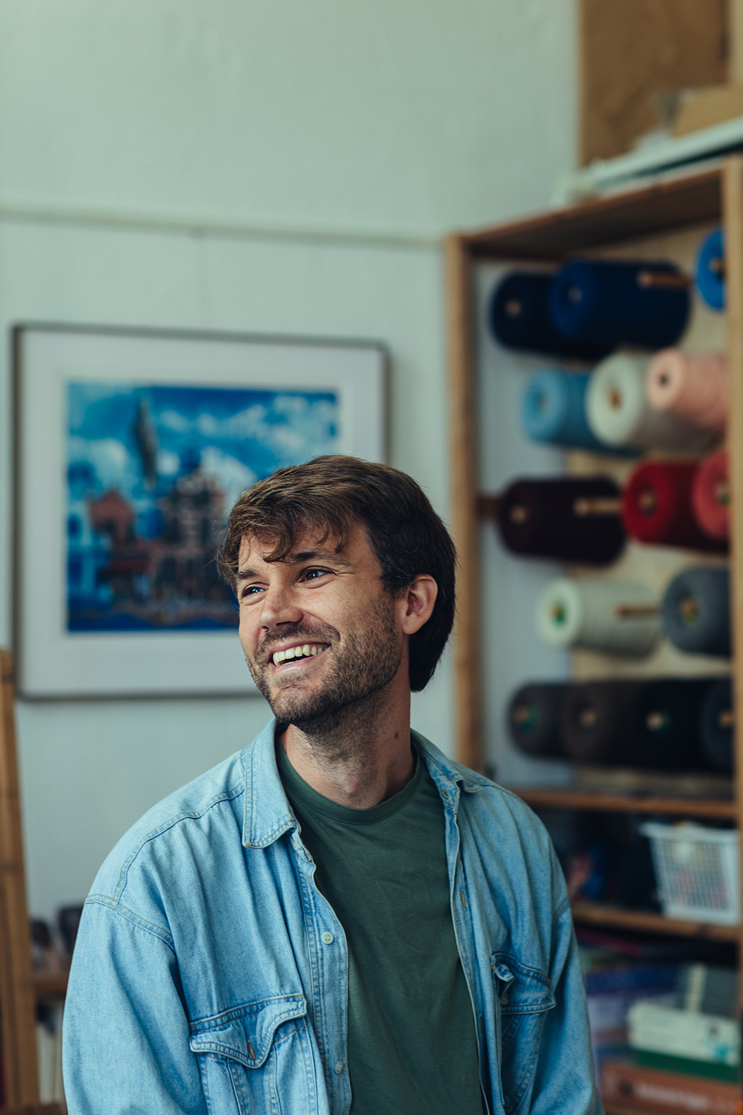 Rafael zit glimlachend in zijn atelier, met een blauw overhemd over een groen t-shirt. Achter hem hangt een blauwdruk schilderij en rollen gekleurd garen, en liggen materialen klaar voor nieuwe creatieve projecten.