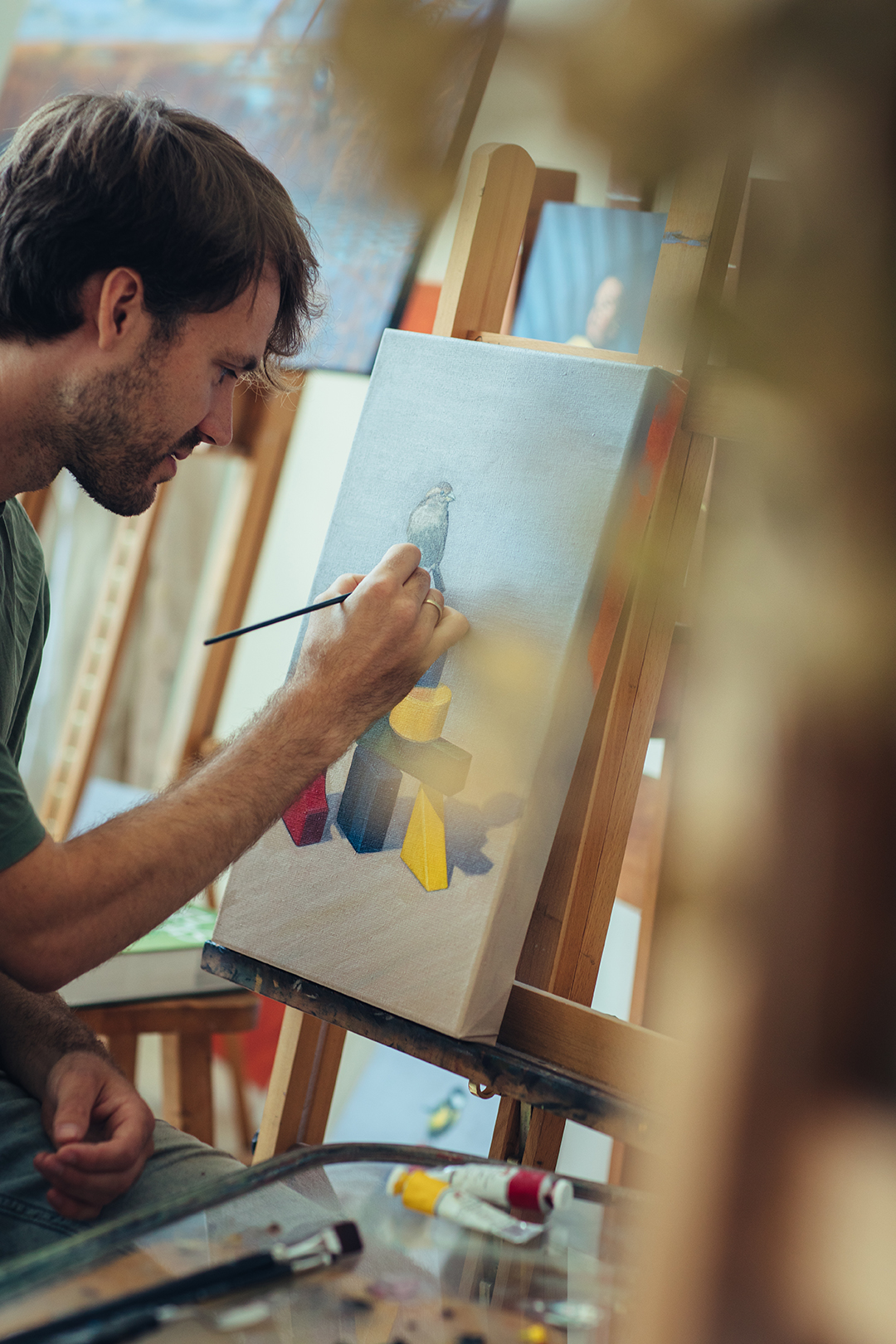 Rafael werkt met een penseel aan een schilderij van een huismus die boven op een constructie van gekleurde houten speelgoedblokken zit. De blauwe, gele, rode en aardse tinten vormen een speels samenspel van geometrische vormen, waartegen de kleine vogel duidelijk afsteekt.
